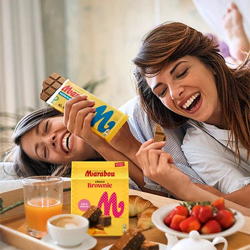 Two smiling women relaxing in bed, sharing Marabou Brownie chocolate bars from a breakfast tray with coffee, strawberries, and pastries.